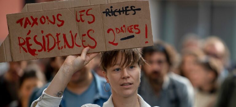 Jeune femme tenant dans les mains une pancarte : Taxons les résidences secondaires en Bretagne