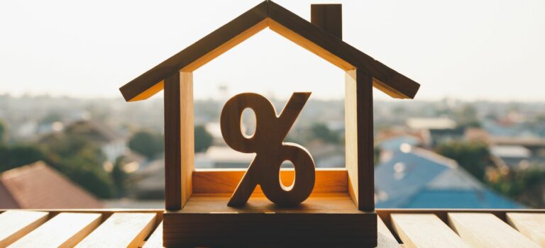 Maison en bois avec un avec taux de crédit à l'intérieur posée sur le rebord d'une fenetre avec la ville en arriere fond pour illustrer la hausse des taux de credits immobiliers et la baisse des prix.