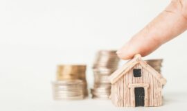 doigt posé sur une maison miniature avec des piles de pièces d'euros en toile de fond pour illustrer les prix de l'immobilier