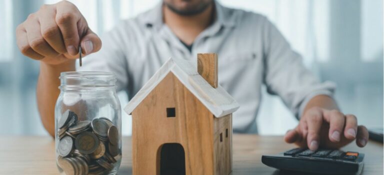 Homme en train de mettre des euros dans un bocal pose sur une table a cote d'une maison miniature