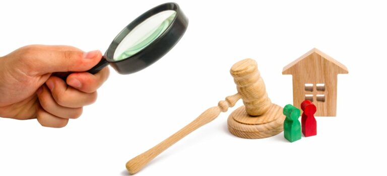 main tenant une loupe examinant une maison, un marteau et deux personnages en bois pour illustrer une question juridique.