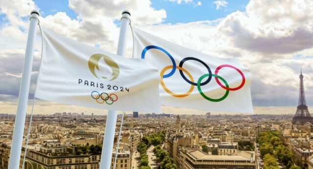 Deux drapeaux flottant avec les logos des Jeux Olympiques de Paris 2024, avec la ville de Paris et la Tour Eiffel en arrière-plan