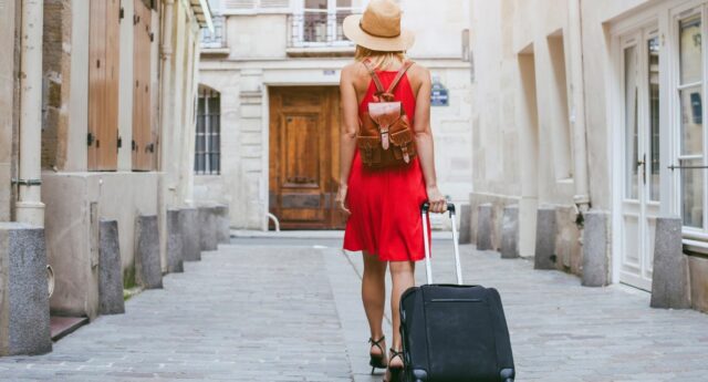 Jeune femme en robe rouge, chapeau de paille et sac a dos en cuir, tirant une valise a roulettes noire, marchant dans une rue pavee etroite et bordee de batiments anciens avec des volets en bois, sous la lumiere du soleil