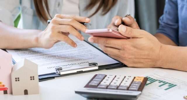 couple un telephone a la main avec une calculatrice posee sur le bureau et des maisons miniatures pour illustrer l'achat immobilier