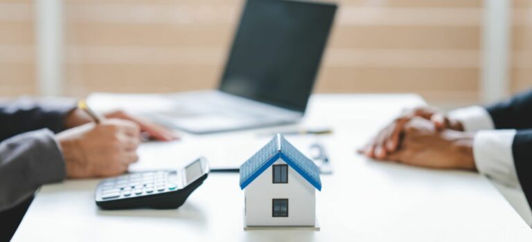 Deux personnes en train de signer un contrat immobilier, avec une petite maison en modele reduit posee sur la table a cote d'une calculatrice et d'un ordinateur portable.