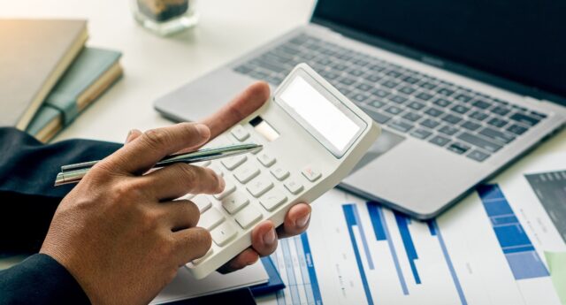 Main tenant une calculatrice tout en consultant des graphiques financiers sur un bureau, avec un ordinateur portable et des documents