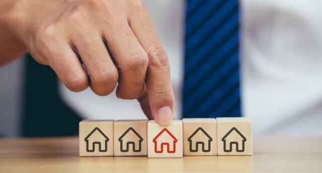 Main d'une personne en costume pointant un cube en bois avec une maison rouge parmi plusieurs cubes avec des maisons noires, symbolisant un choix immobilier