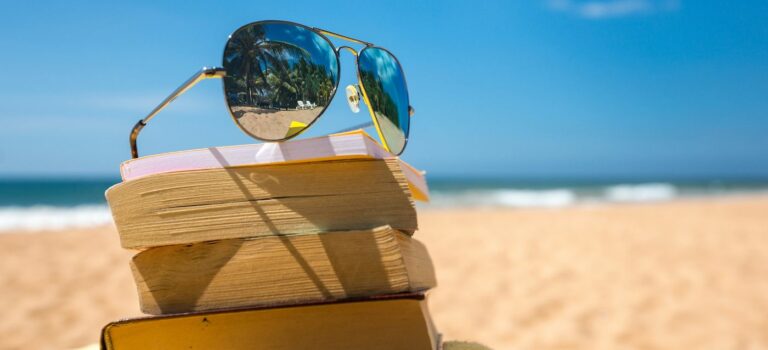 des lunettes de soleil posees sur une pile de livres sur une plage avec la mer au fond