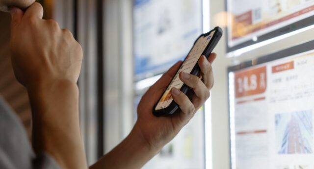 Une personne consulte des annonces immobilieres sur son telephone devant une vitrine d’agence.