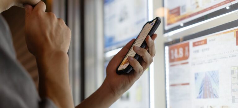 Une personne consulte des annonces immobilieres sur son telephone devant une vitrine d’agence.