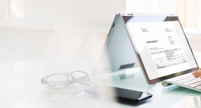 Professionnel saisissant des factures sur un ordinateur portable, entouré de documents comptables et d'une calculatrice.