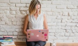 Femme utilisant un ordinateur portable rose, assise devant un mur de briques blanches.