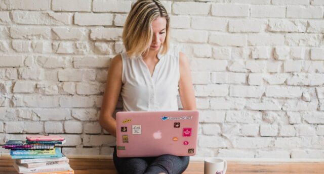 Femme utilisant un ordinateur portable rose, assise devant un mur de briques blanches.