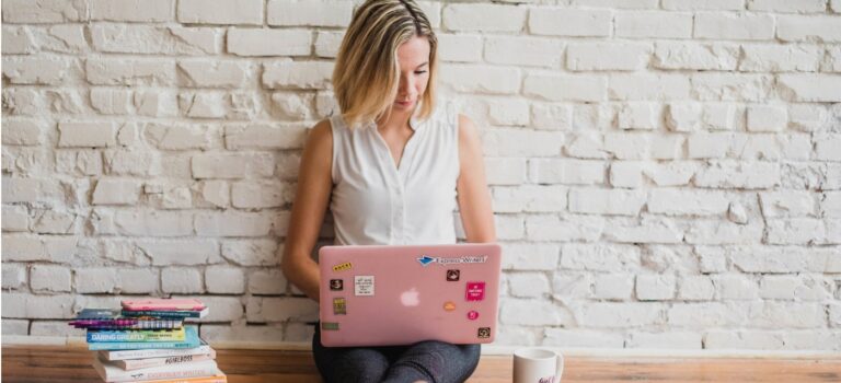 Femme utilisant un ordinateur portable rose, assise devant un mur de briques blanches.