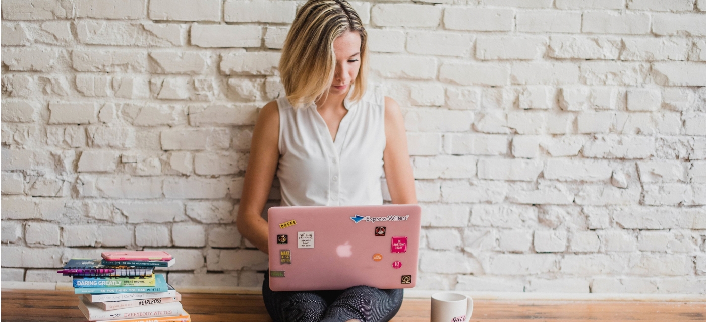 Femme utilisant un ordinateur portable rose, assise devant un mur de briques blanches.