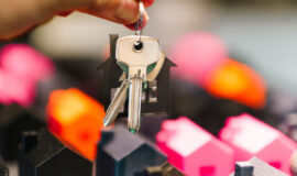 Une main tient un trousseau de clés avec un porte-clés en forme de maison devant des maisons miniatures colorées.