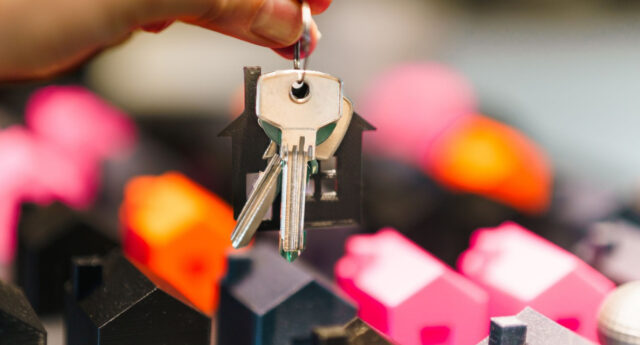 Une main tient un trousseau de clés avec un porte-clés en forme de maison devant des maisons miniatures colorées.