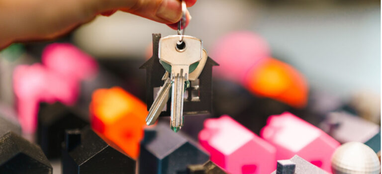 Une main tient un trousseau de clés avec un porte-clés en forme de maison devant des maisons miniatures colorées.